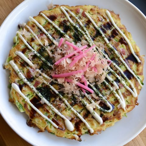 Golden-brown Japanese Okonomiyaki Pancakes sizzling on a griddle, garnished with vibrant green aonori and pickled ginger for a savory bite.  