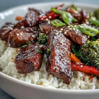 Sizzling teriyaki beef slices glisten over fluffy white rice with vibrant sautéed broccoli and peppers.  